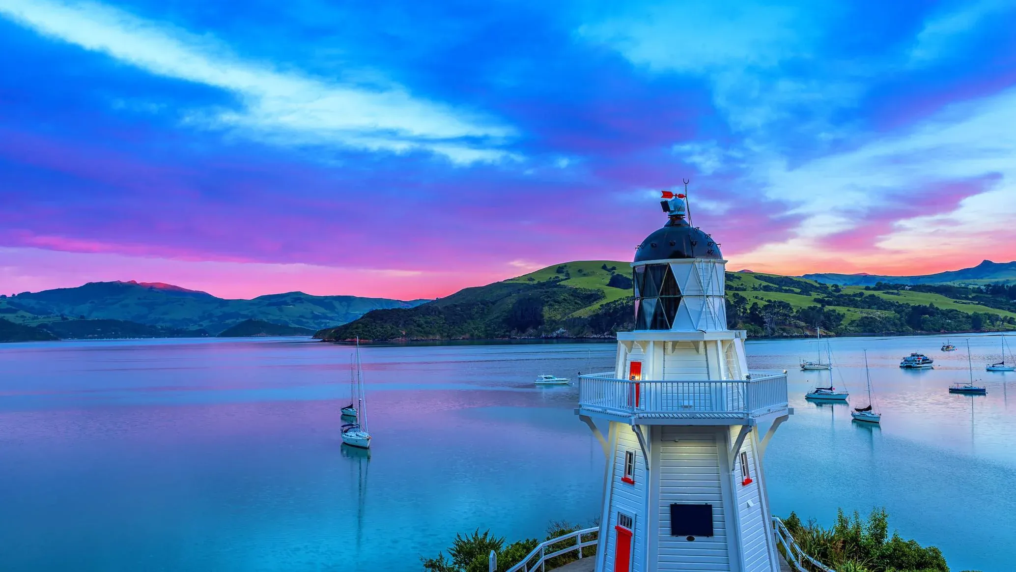 Scenic Akaroa lighthouse at sunset, overlooking serene waters and yachts against a vibrant sky in New Zealand.