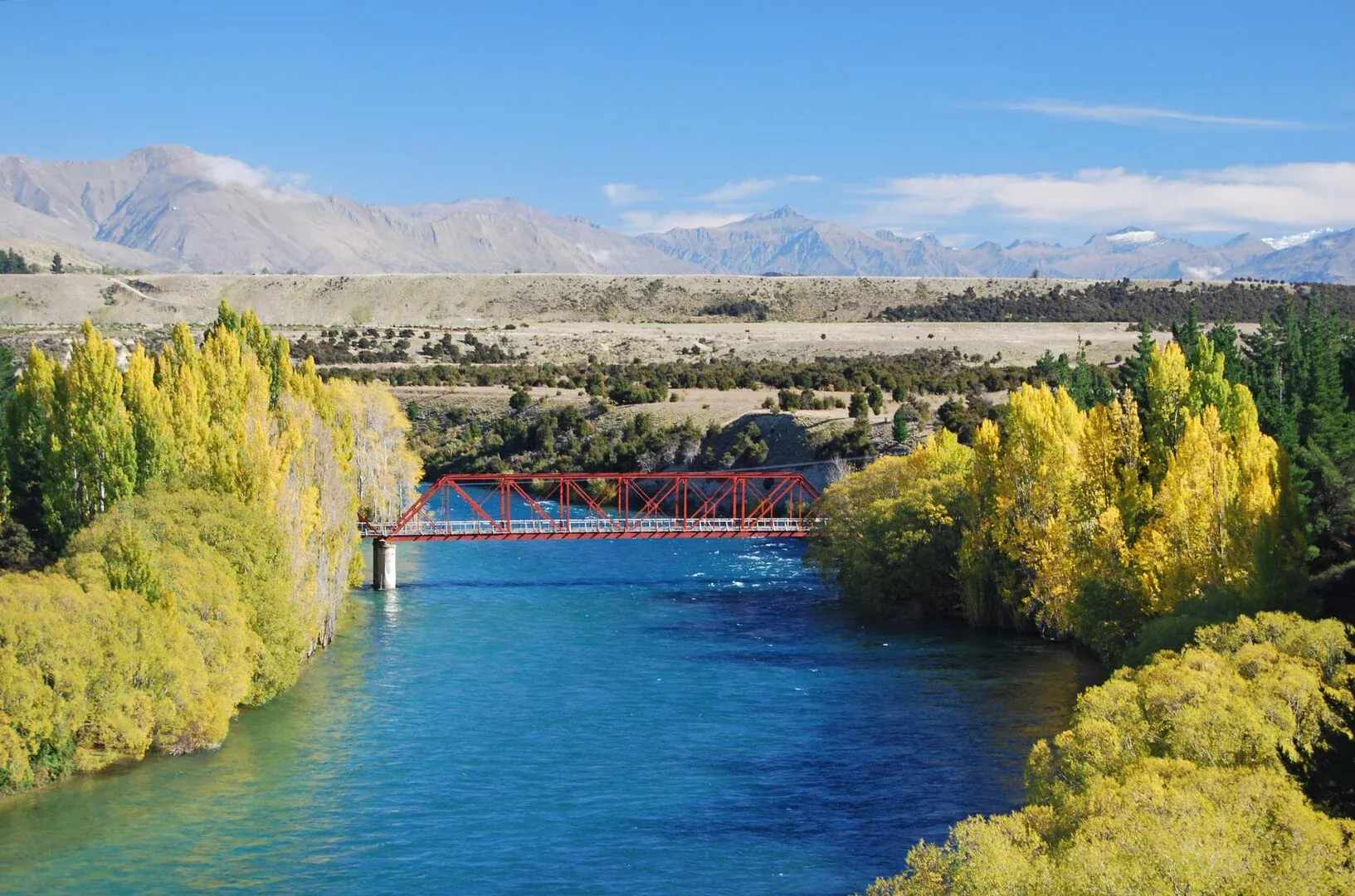 Red bridge over Clutha River, surrounded by autumn foliage in Luggate, Otago, New Zealand.